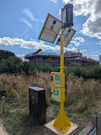 The wind and solar powered defib cabinet in the nature corridor at Houlton with construction of houses being undertaken nearby