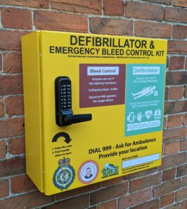Yellow metal locked defibrillator and bleed kit cabinet, mounted on a brick wall. 